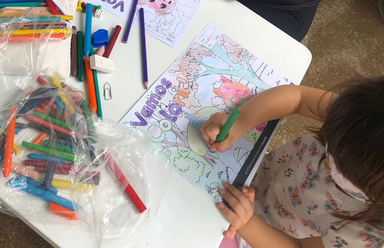 Na imagem há uma criança sentada em uma mesa, pintando com giz de cera um desenho que tem um gato e um cachorro com os dizeres "Vamos colorir!".