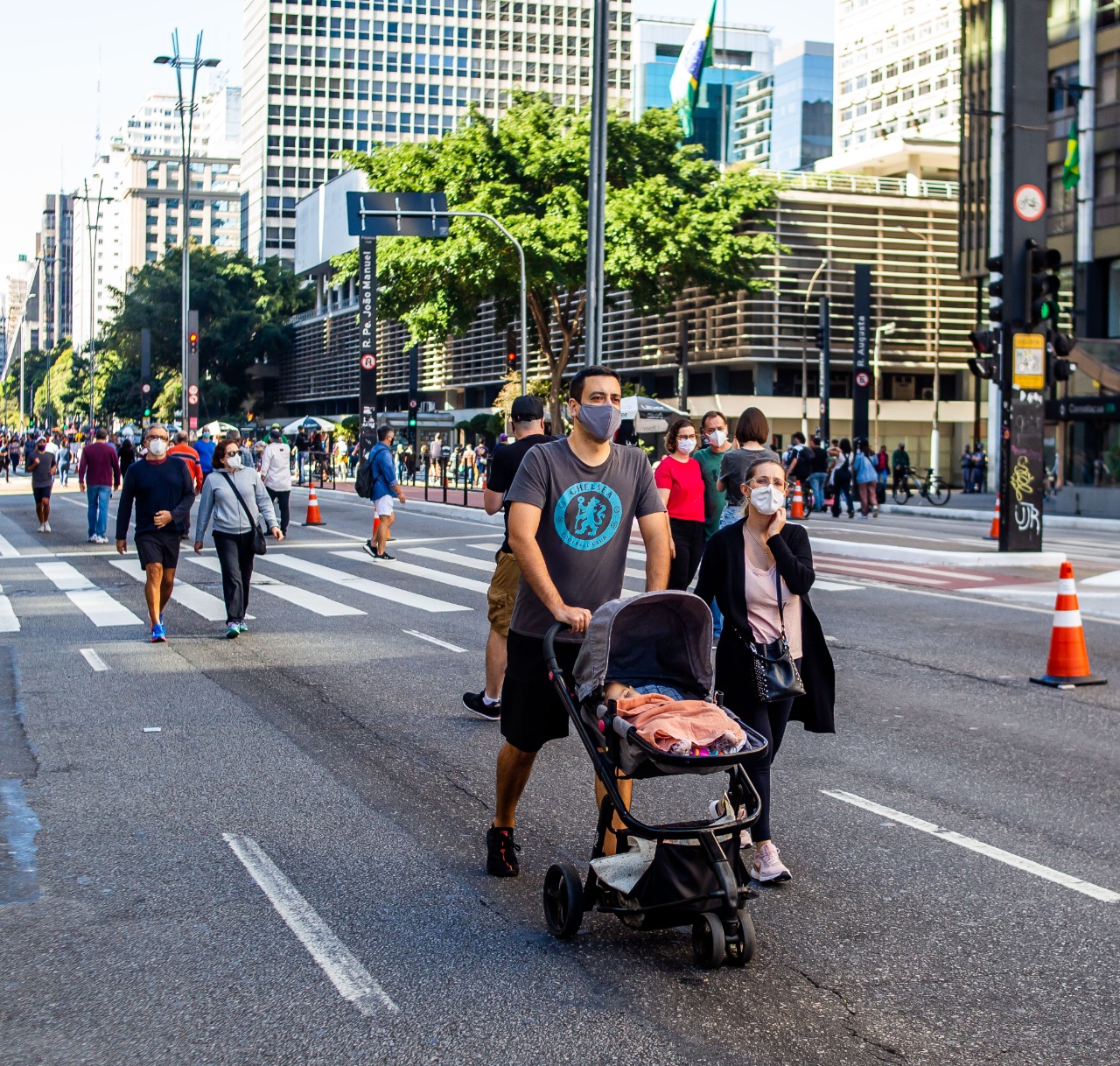 Foto de movimentação na via. Pessoas de máscara e trajes esportivos passeiam na pista da avenida Paulista. Em primeiro plano, um casal empurra um carrinho de bebê. Ao fundo temos edifícios altos