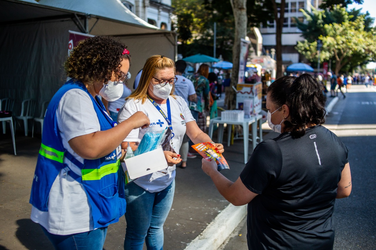 Foto de abordagem. Duas agentes de Saúde uniformizadas e de máscara conversam com uma mulher que caminhava pela pista aberta da avenida Paulista. Uma delas entrega um panfleto. Ao fundo temos a movimentação nos estandes