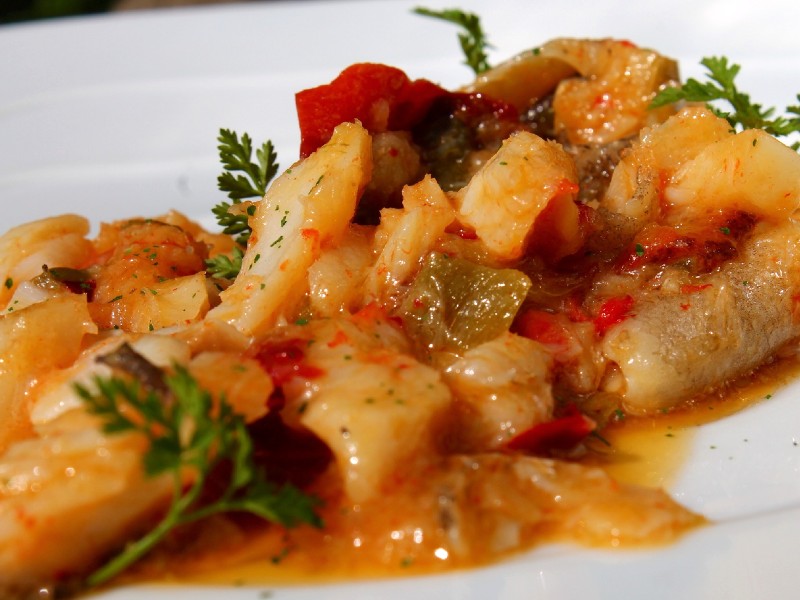 Foto de um prato com bacalhau. O peixe está cozido com molho e legumes como pimentão e batata e está disposto num prato branco, com folhas verdes de salsinha na decoração