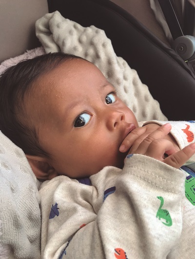 Num carrinho e de roupa branca, um bebê negro de olhos escuros está em close, com as mãos na boca, olhando para a câmera.