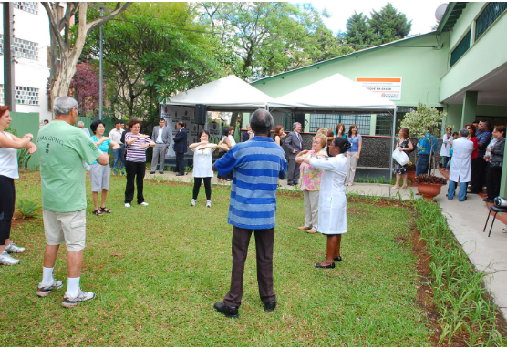 Há várias pessoas fazendo atividade de Prática Integrativa, com orientação de uma profissional da saúde, na área externa do Centro de Referência em Homeopatia Medicina Tradicional e Práticas Integrativas em Saúde