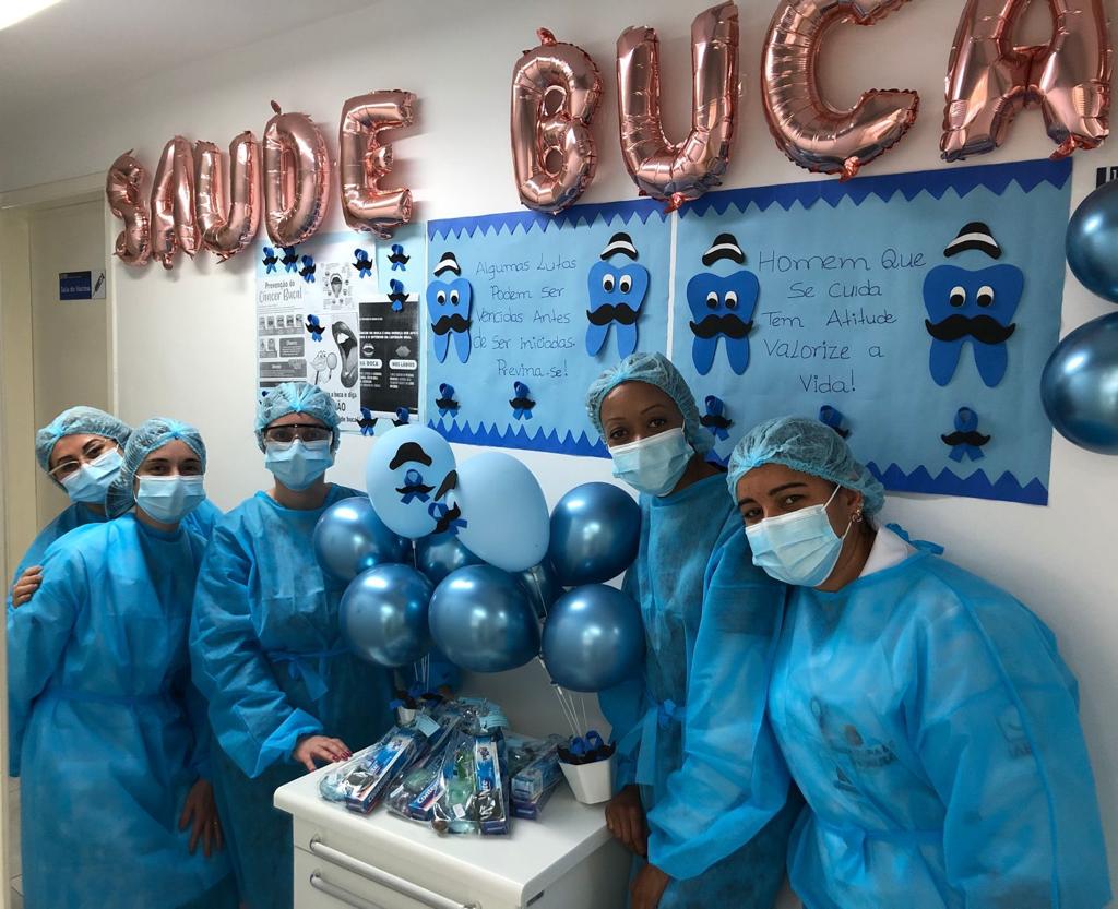 Na imagem há cinco profissionais da saúde usando roupas de proteção, máscara e touca azuis. Todos estão ao lado de uma mesa branca com bexigas e sacolas azuis. Ao fundo há uma parede escrito Saúde Bucal com balões e cartolinas azuis com desenhos de dentes.