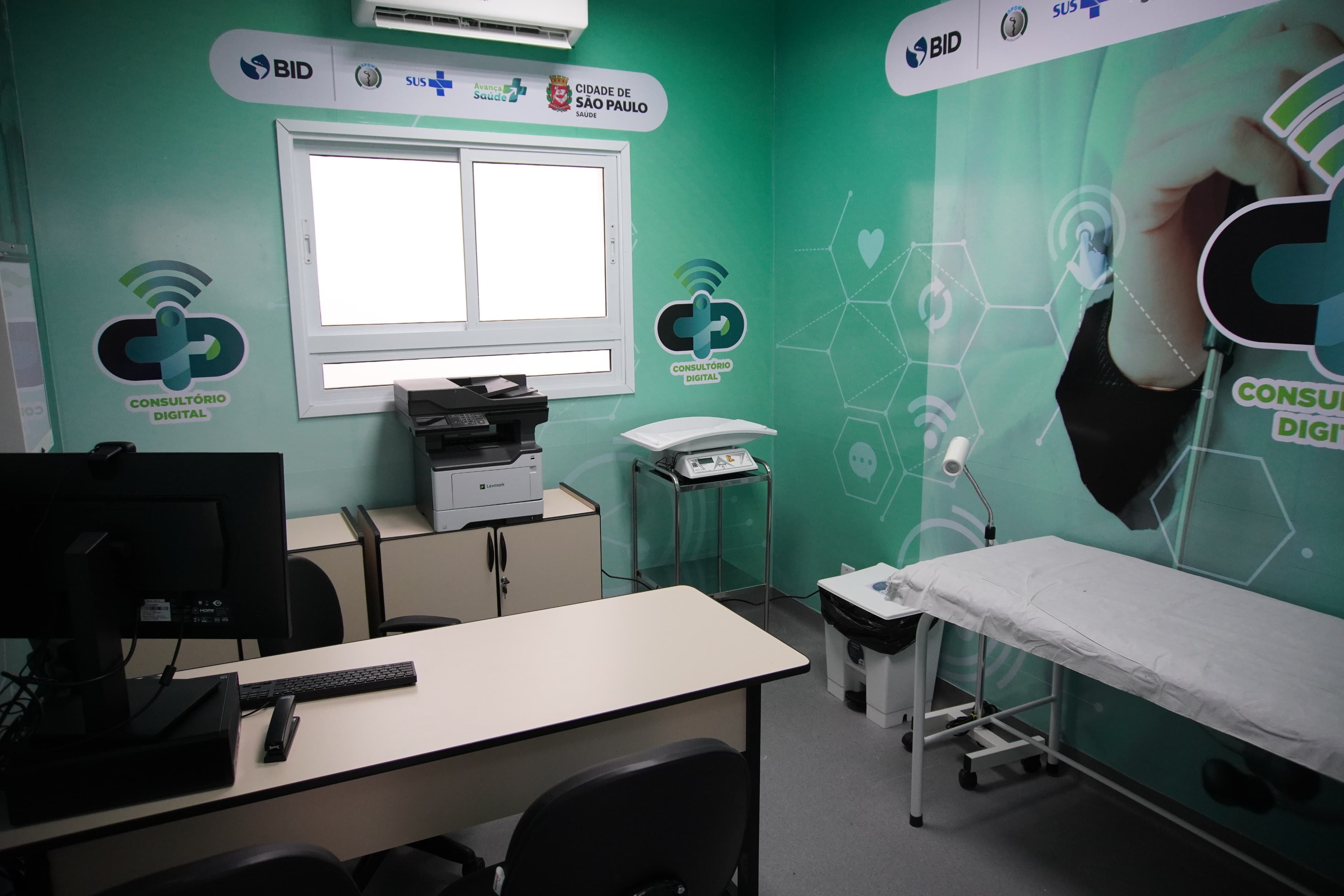 Imagem mostra uma sala da UBS equipada com mesa, computador, impressora, maca e decorada com temas de saúde