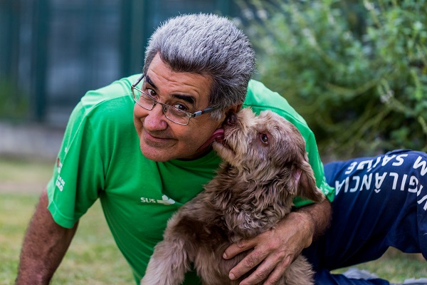 #PraCegoVer: um agente de vigilância em saúde está num local aberto, sentado na grama olhando para quem o fotografa enquanto um cachorro de pelo longo, cor marrom claro, lambe o rosto dele.