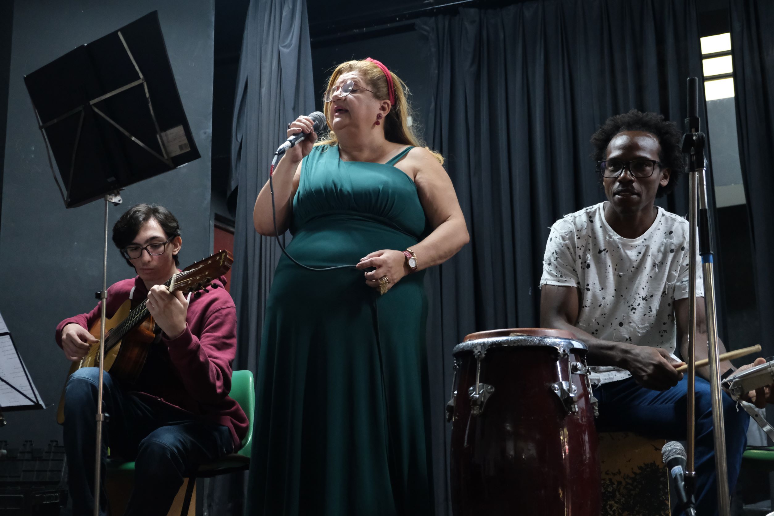 Na imagem, da esquerda para a direita, está Pedro Puccini sentado tocando violão, enquanto ao seu lado Nádia Maria canta. Logo em seguida está Heverton tocando percussão