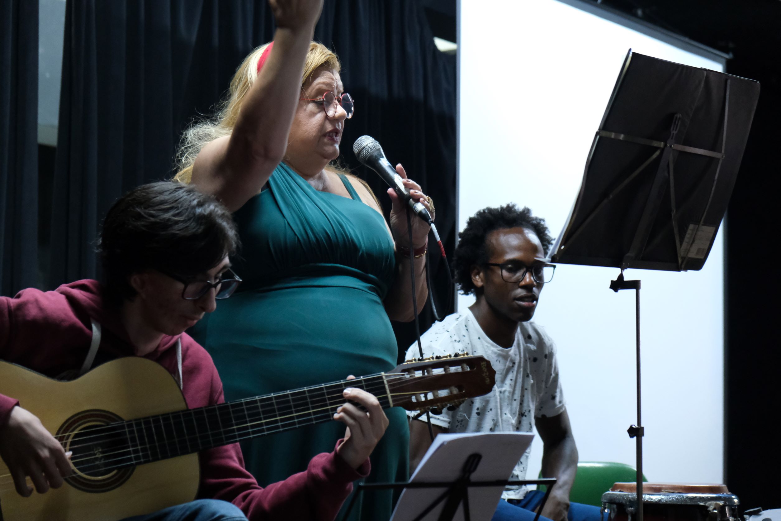 Na imagem, da esquerda para a direita, está Pedro Puccini sentado tocando violão, enquanto ao seu lado Nádia Maria canta. Logo em seguida está Heverton tocando percussão