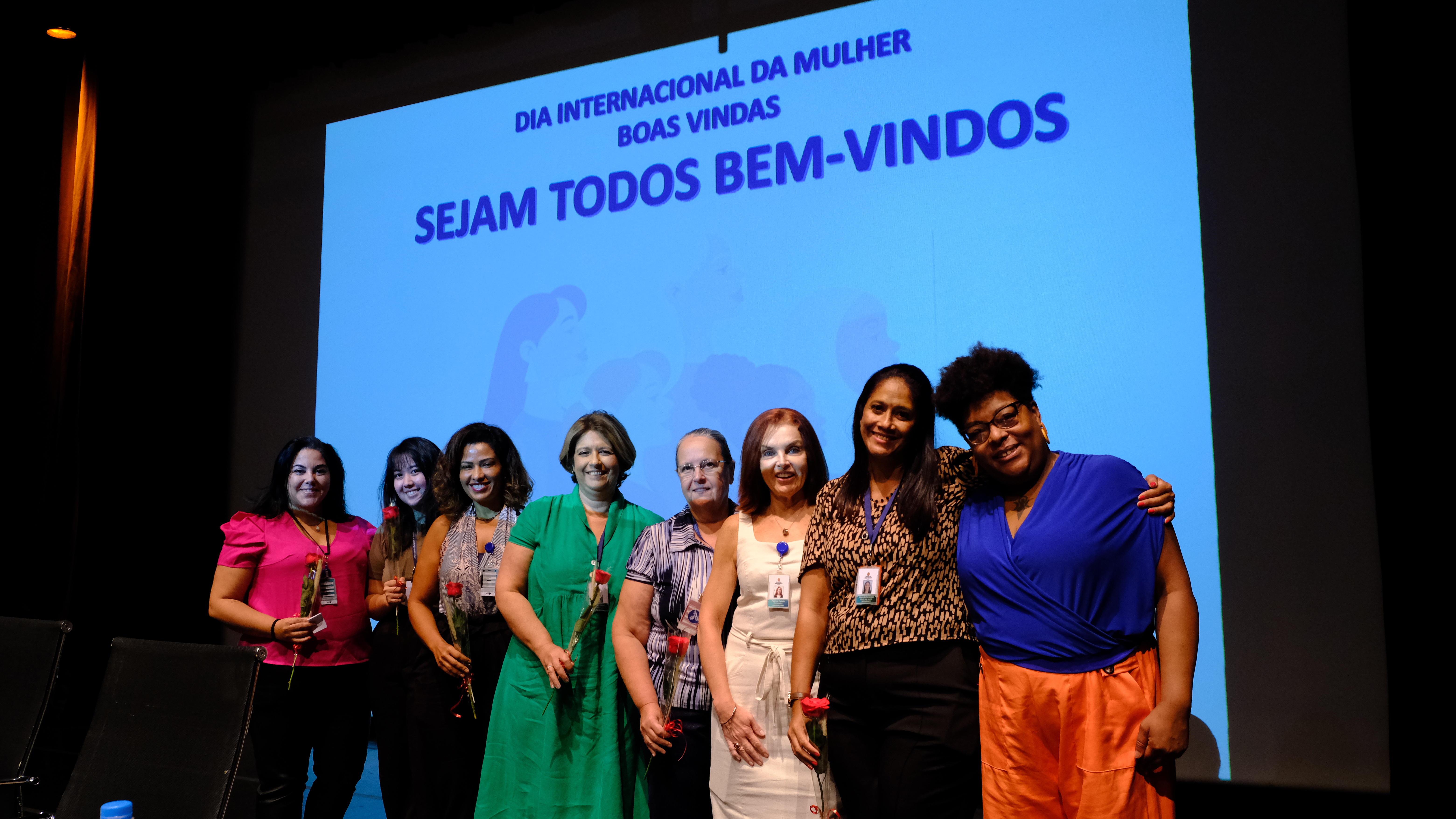 Na imagem estão oito mulheres com uma rosa na mão, uma ao lado da outra sorrindo para a foto. Todas participaram do evento. 