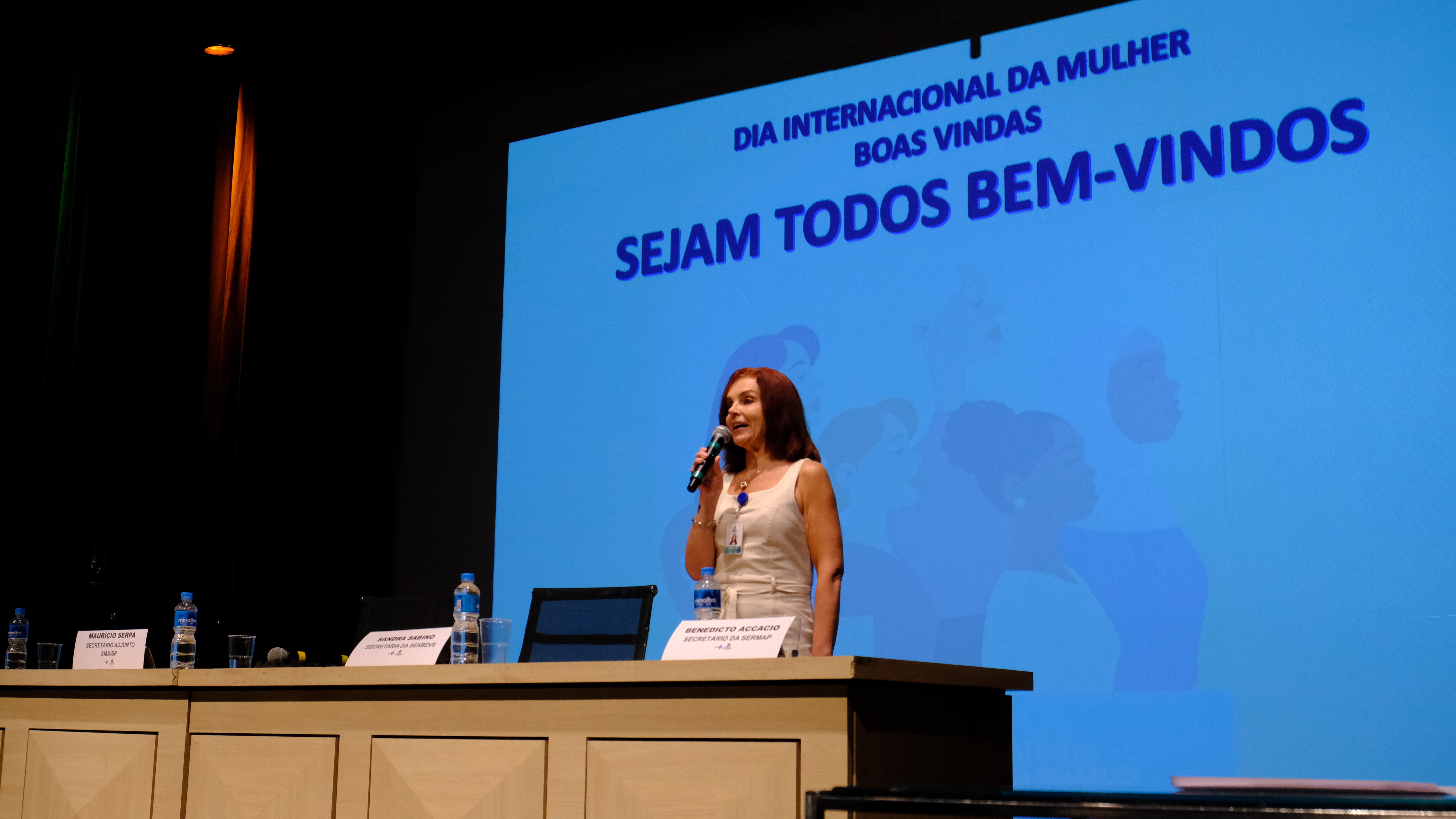 A Coordenadora de Gestão de Pessoas, Patrícia Pallota, está em pé no centro da imagem falando ao público do evento