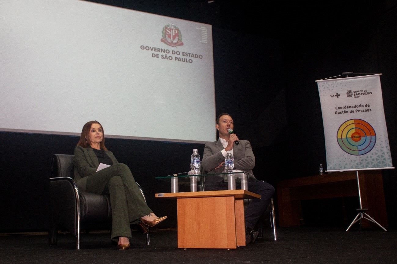 Na imagem, à esquerda, está Patrícia Pallota sentada em uma cadeira , ao seu lado direito, também sentado, está o Secretário Adjunto da SMS, Dr. Maurício Serpa falando ao microfone que está em sua mão