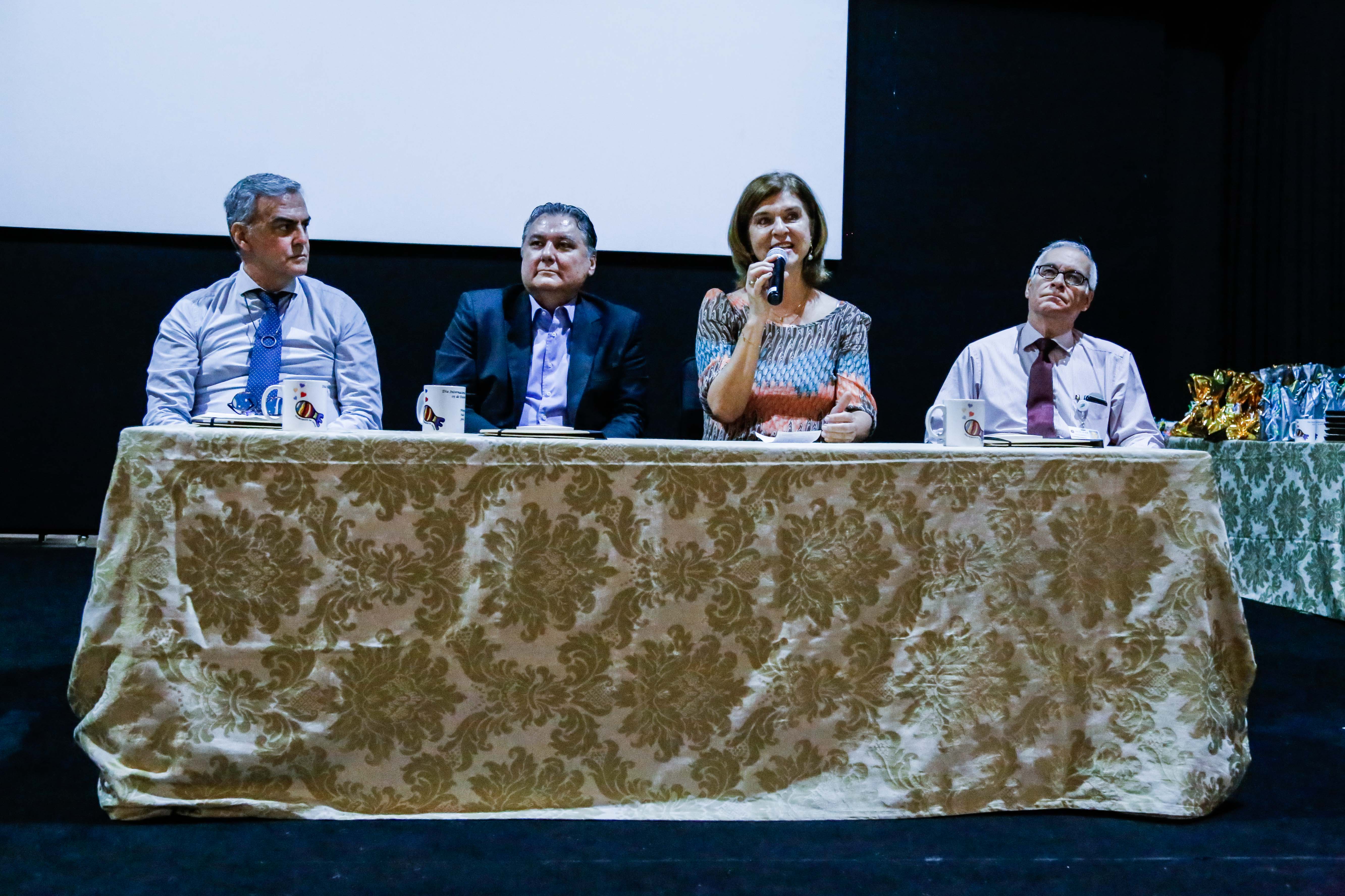 Quatro pessoas estão sentadas a uma mesa montada em cima do palco do teatro. A coordenadora Jane Abrão, está sentada e é a terceira da esquerda para direita, está com o microfone na mão.
