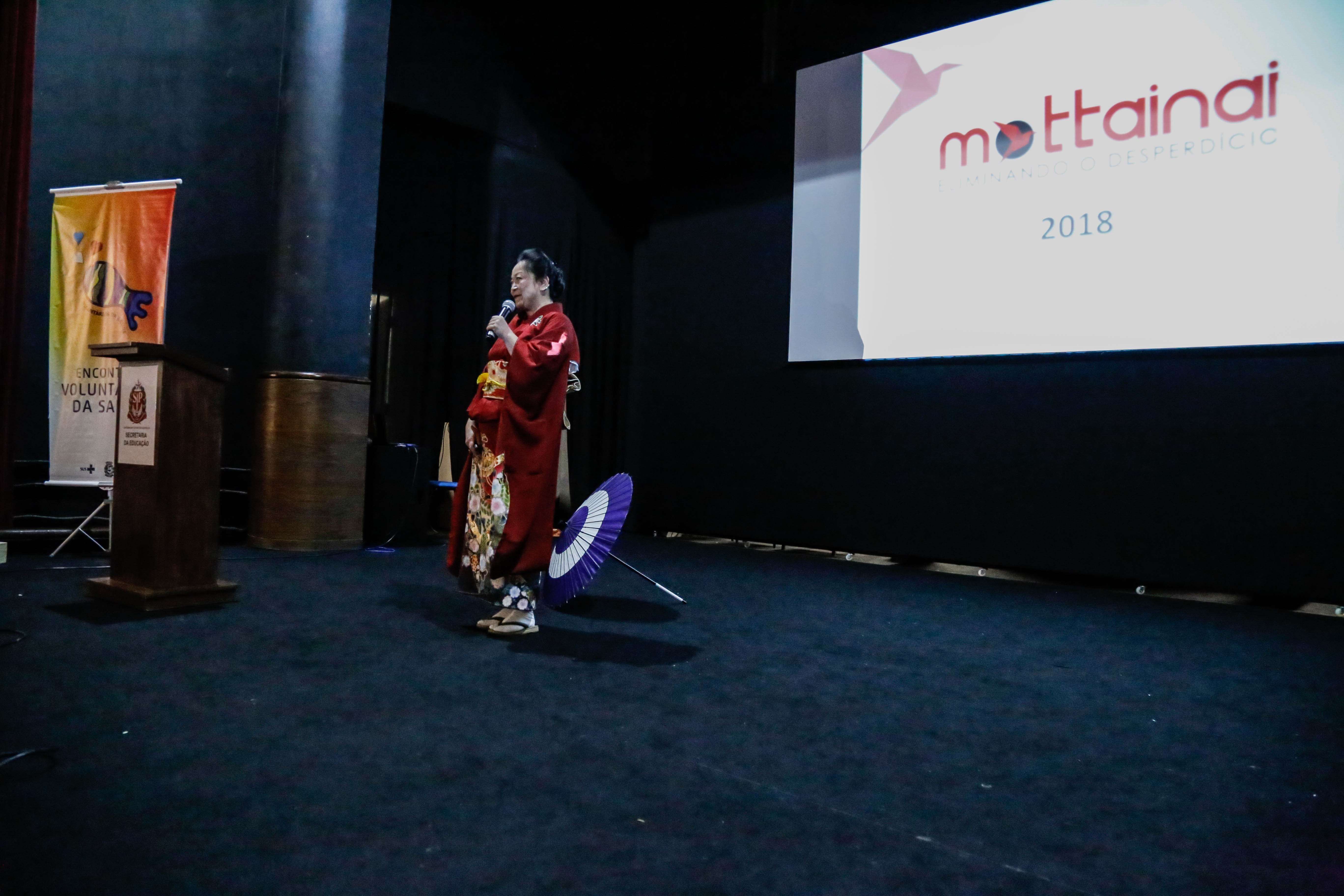 Lilian Tiemi Yamashita está no meio do palco fazendo sua palestra. Ao seu lado, um guarda-chuva colorido está no chão. No fundo do palco está a palavra Mottainai no telão.