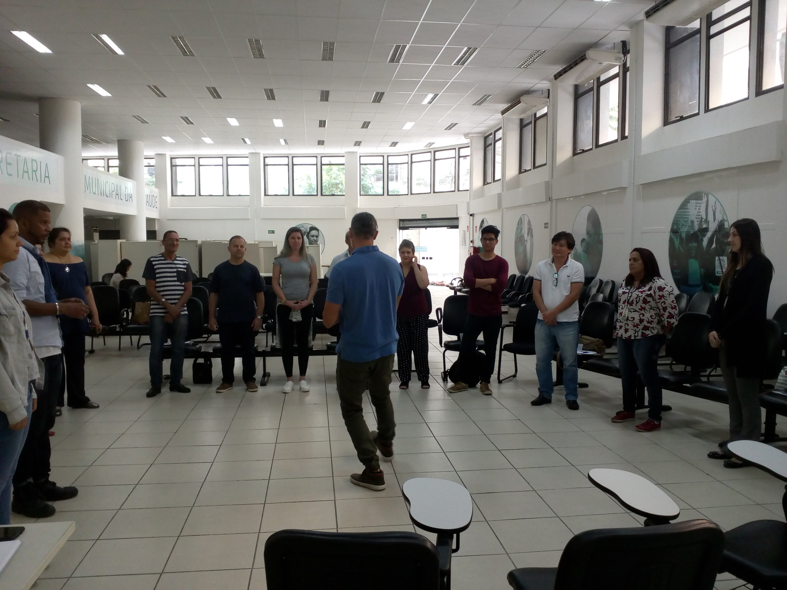 #Paracegover Foto frontal: No centro da imagem, se encontra o palestrante de camisa azul que está de costas para a foto ensinando a atividade para os participantes. que estão em pé na frente do palestrante formando um semicírculo em volta dele