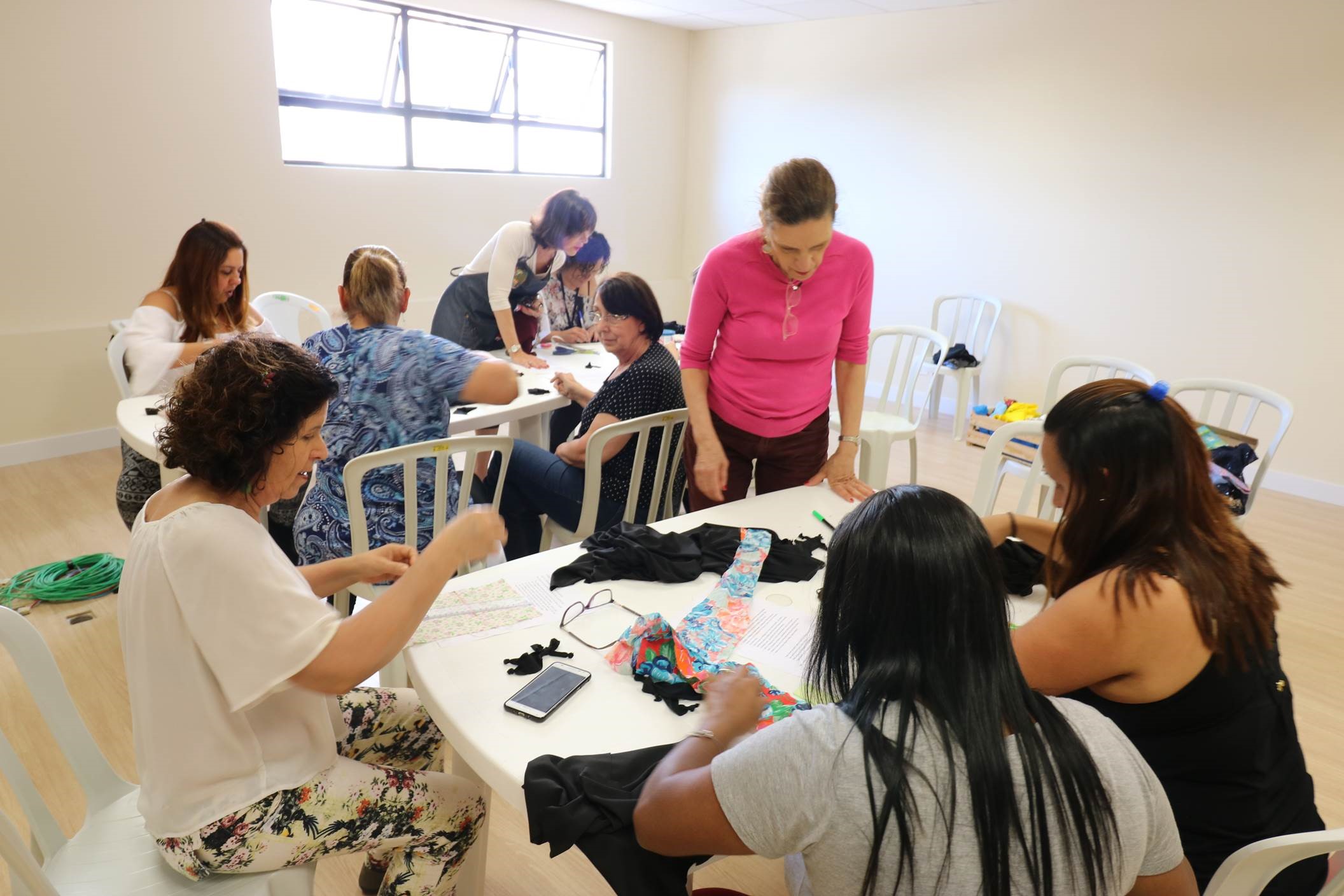 #Paracegover Na imagem estão dois grupos de mulheres. Cada grupo está sentado em uma mesa criando bonecas.