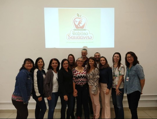 #Paracegover Na imagem estão os participantes do evento juntamente com os organizadores do evento, posando para foto, acima deles está projetado o logo do hábitos saudáveis, uma maçã com um homem dentro dela praticando exercícios físicos, escrito Hábitos Saudáveis abaixo da maçã.