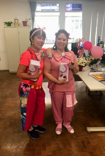 #Paracegover Na imagem estão as duas voluntárias vestidas de palhacinhas, em pé e ambas vestidas toda de rosa. Na mesa de escritório ao lado delas estão um vaso de flores e três balões rosas.