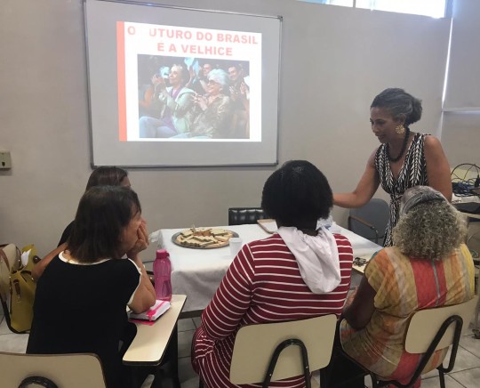 #Paracegover Na imagem estão sentadas quatro servidoras ao redor de uma mesa. A instrutora está em pé, falando com elas. Trata-se de uma atividade em grupo realizada no evento.