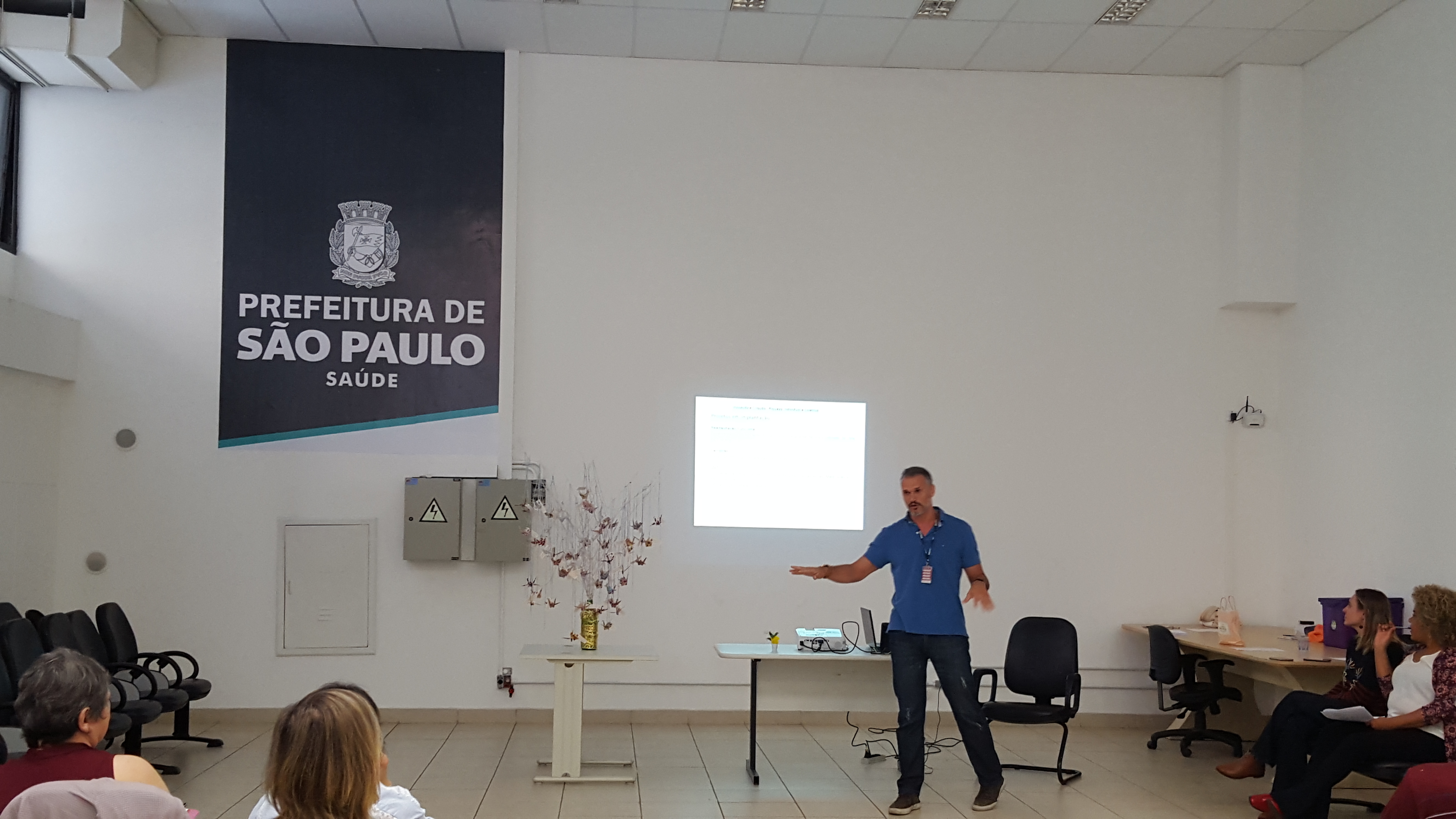 #Paracegover Na imagem: Um servidor está no centro da imagem falando sobre seu projeto. Atrás dele está uma imagem com o logo da Prefeitura Municipal de São Paulo.