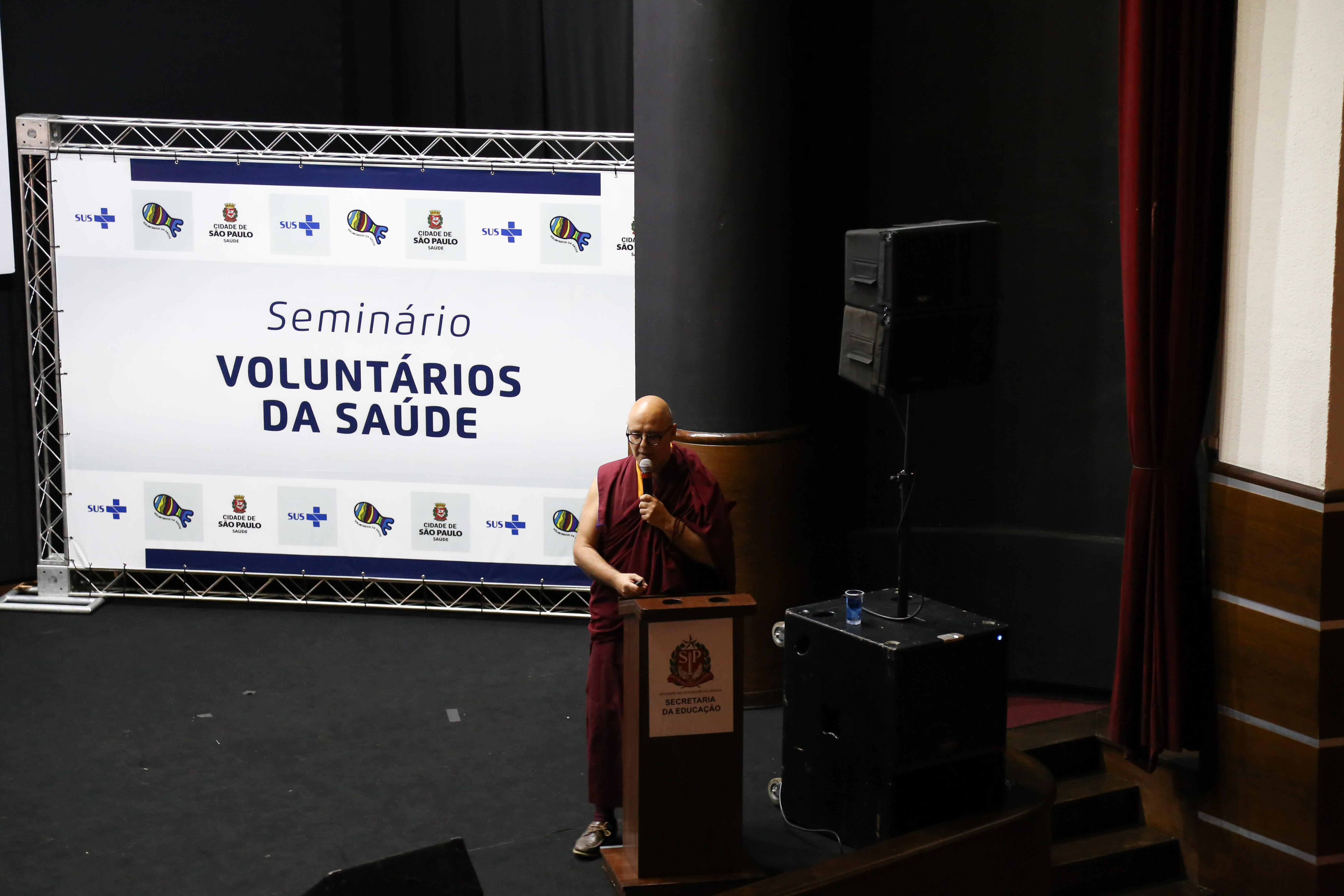 #Paracegover Na imagem está o monge em cima do palco conversando com o público, atrás dele está um banner escrito Seminário Voluntários da Saúde