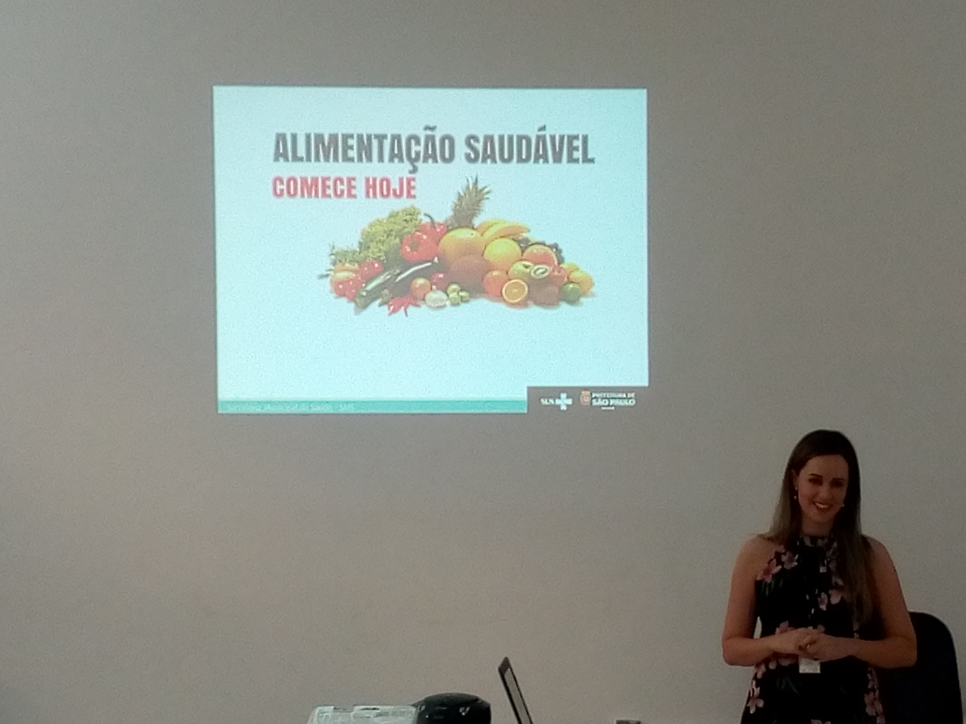 #Paracegover Na imagem; ao fundo, na parede, um projetor reflete um Slide que está escrito Alimentação Saudável Comece hoje, ao lado do slide está a palestrante.