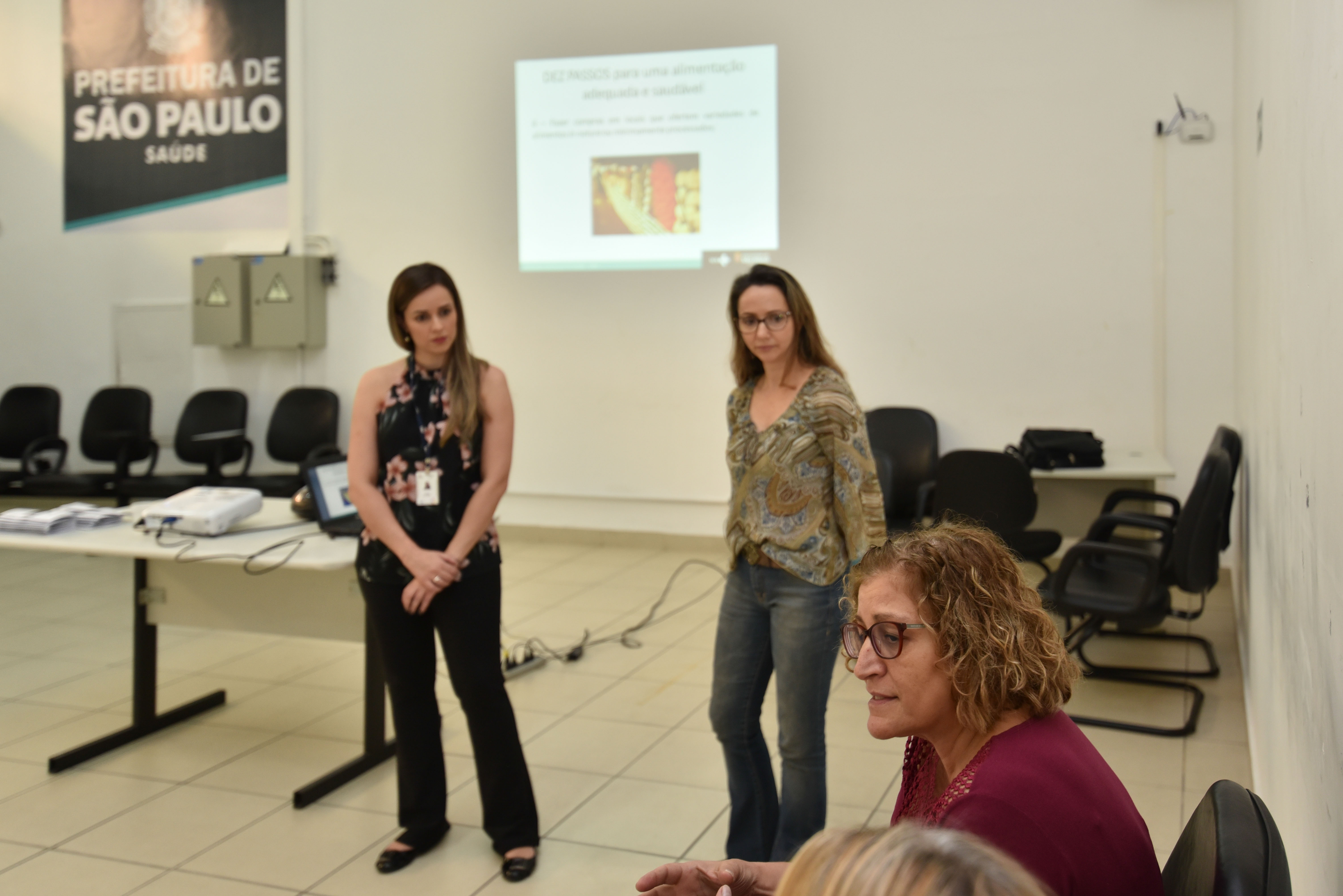 #Paracegover Na imagem: as nutricionistas Mariana Delega de Souza e Daniela Wenzel estão em pé ouvindo uma servidora que sentada à frente delas, no canto direito da foto 
