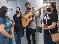 Quatro pessoas estão na foto. Duas mulheres à esquerda, logo após um rapaz com um violão na mão, e por último, no canto direito, a última mulher.
