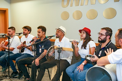 Na imagem está o grupo de samba formado por sete integrantes. Todos estão sentados um ao lado do outro com seus instrumentos