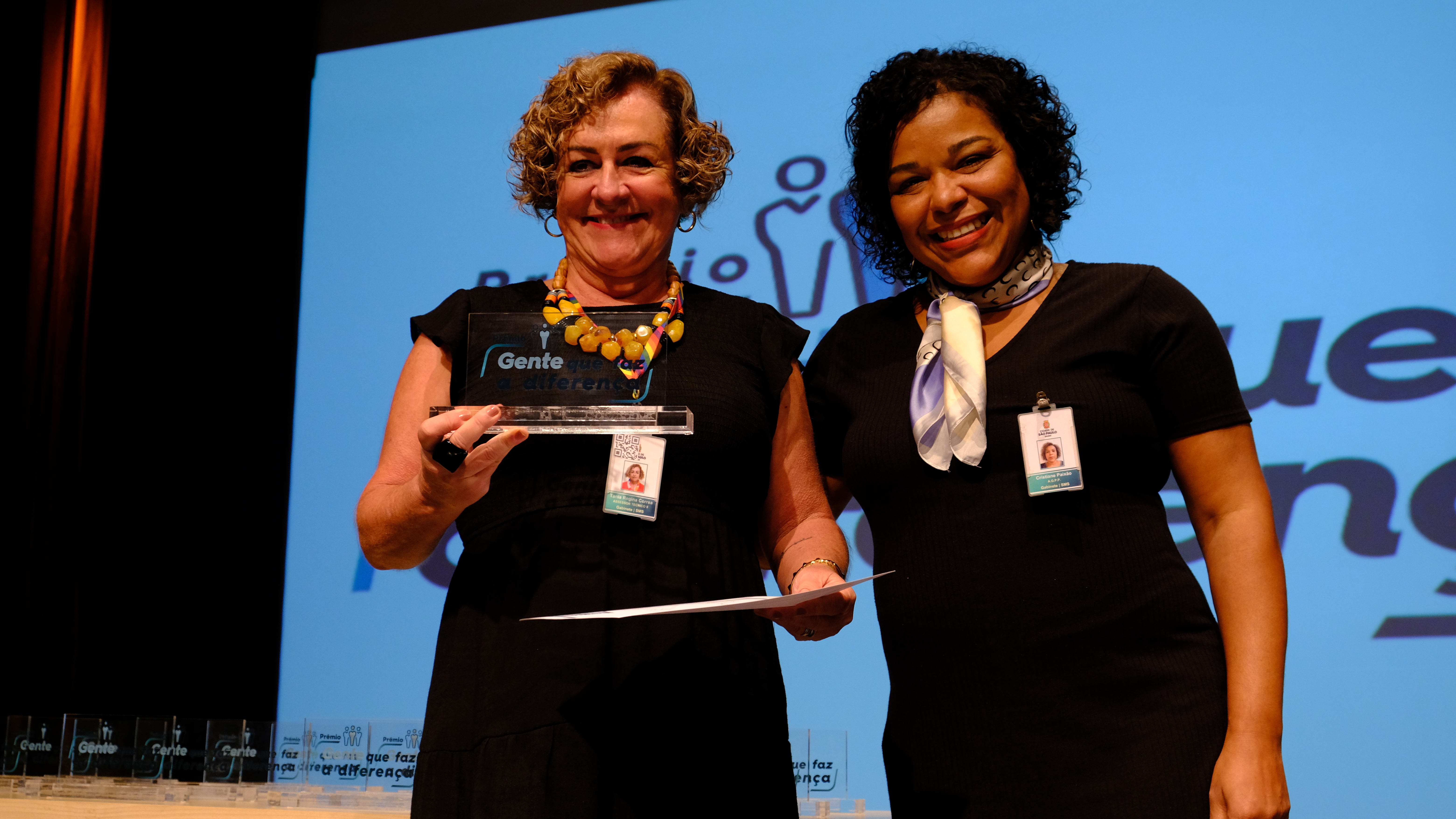 Na imagem está Cristiane Paixão entregando o prêmio para uma das vencedoras