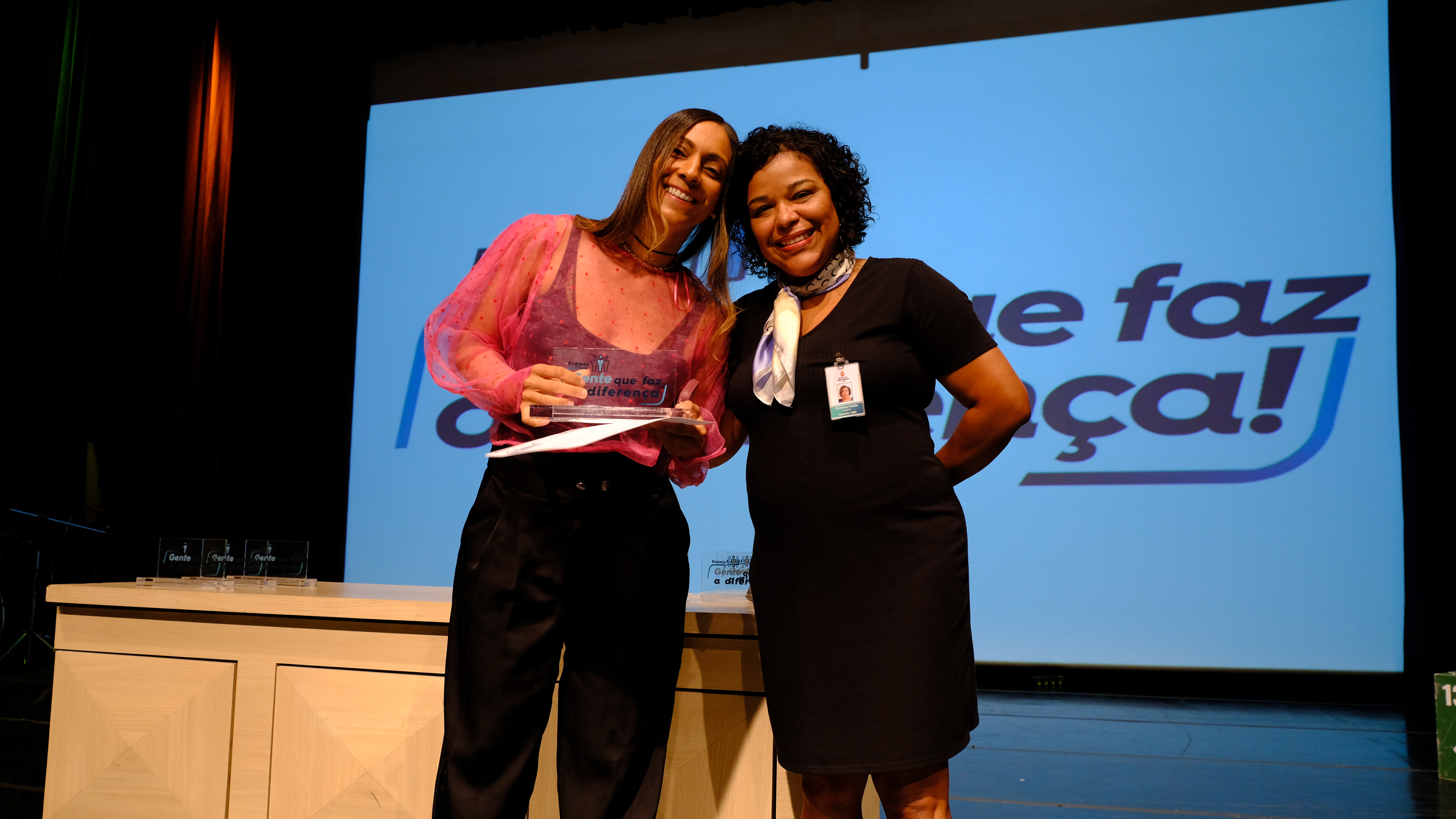 Na imagem está Cristiane Paixão entregando o prêmio para uma das vencedoras