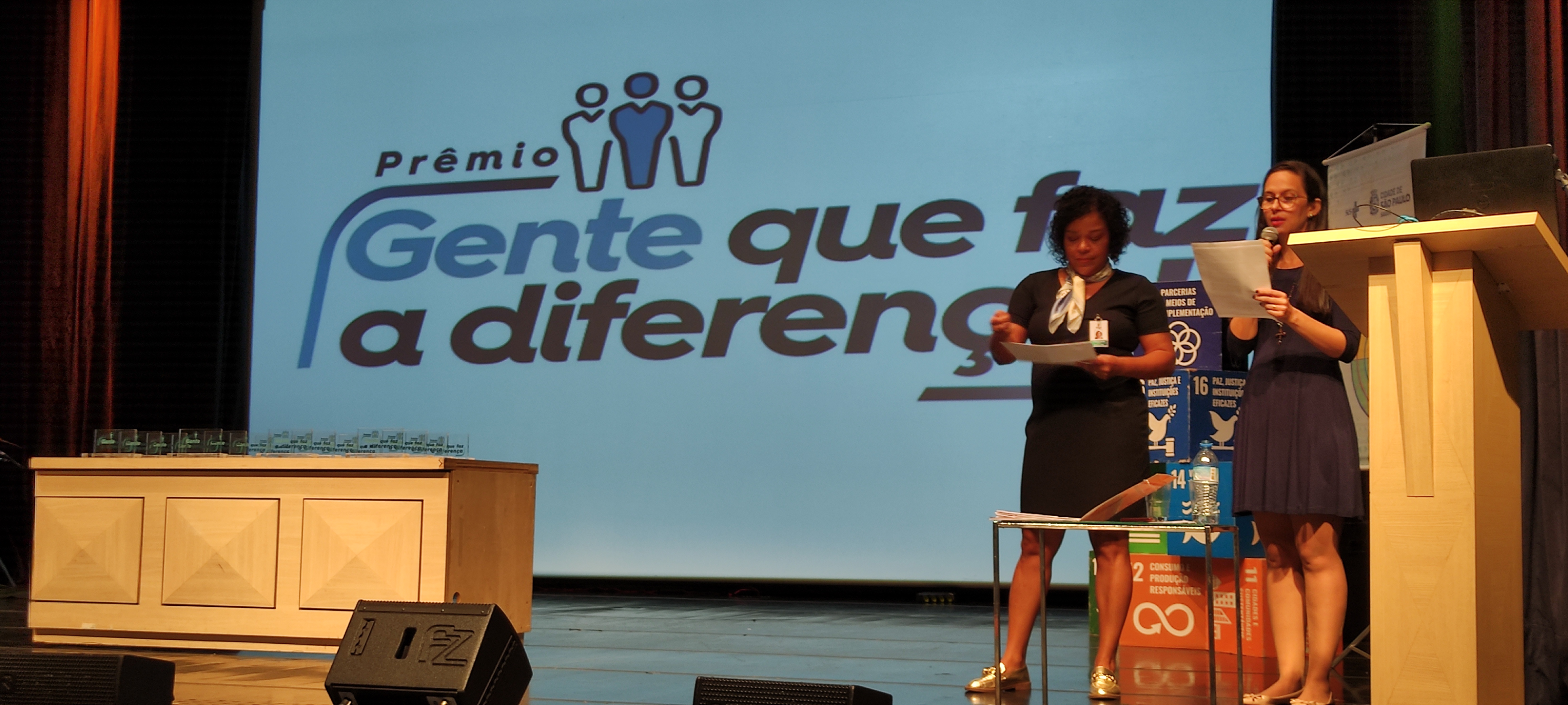No canto direito da imagem está Moema Moraes do Valle, ao seu lado esquerdo está Cristiane Paixão, diretora do DDCQVT. Ao fundo está uma projeção escrito Gente que faz a diferença