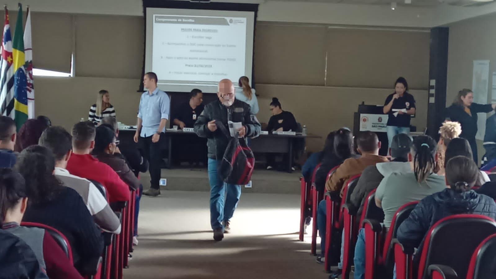 Na imagem há um auditório. Os candidatos estão divididos em dois lados. No meio, há um corredor, um homem caminha por ele após ter escolhido sua vaga. 