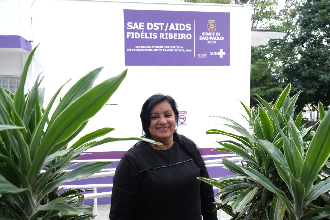 Entre duas plantas verdes, está uma mulher de cabelos pretos, vestindo uma blusa de frio preta com uma listra cinza acima do peito. Ela está sorrindo. Atrás dela, está uma placa roxa com "SAE DST/Aids Fidélis Ribeiro". 