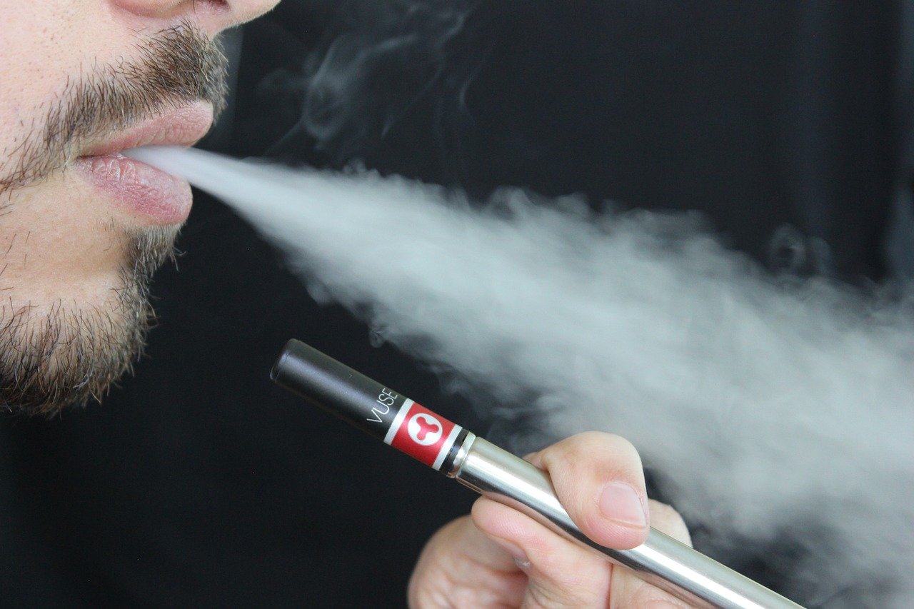 Foto de fumo. Um homem com barba e bigode está soltando fumaça branca pela boca. Em frente à sua boca, ele segura um cigarro eletrônico em formato de canudo entre o dedão e o dedo indicador.