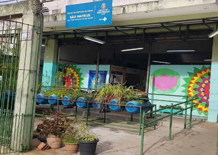 Imagem da fachada do Centro de Práticas Naturais (CPN) São Mateus. Há duas paredes com desenhos coloridos, barris reciclados com plantas e um portão verde.