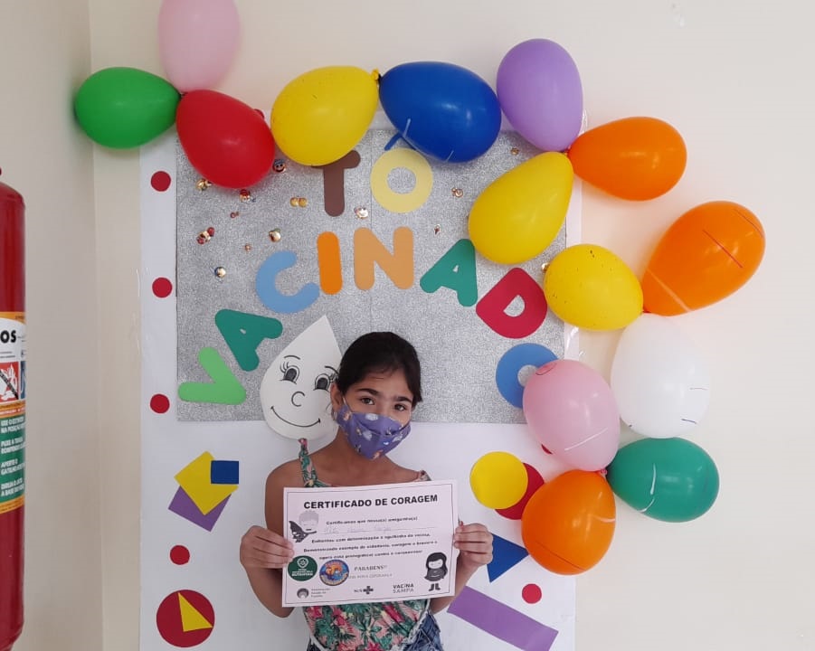 Em uma parede branca estão colados balões coloridos. Atrás deles, está uma placa cinza com letras coloridas 