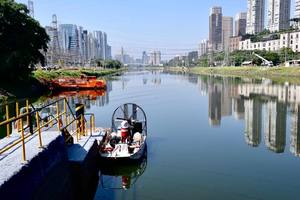 Foto de paisagem. Em um rio com água escura está um barco vazio, ancorado em uma doca com grade amarela e preta.