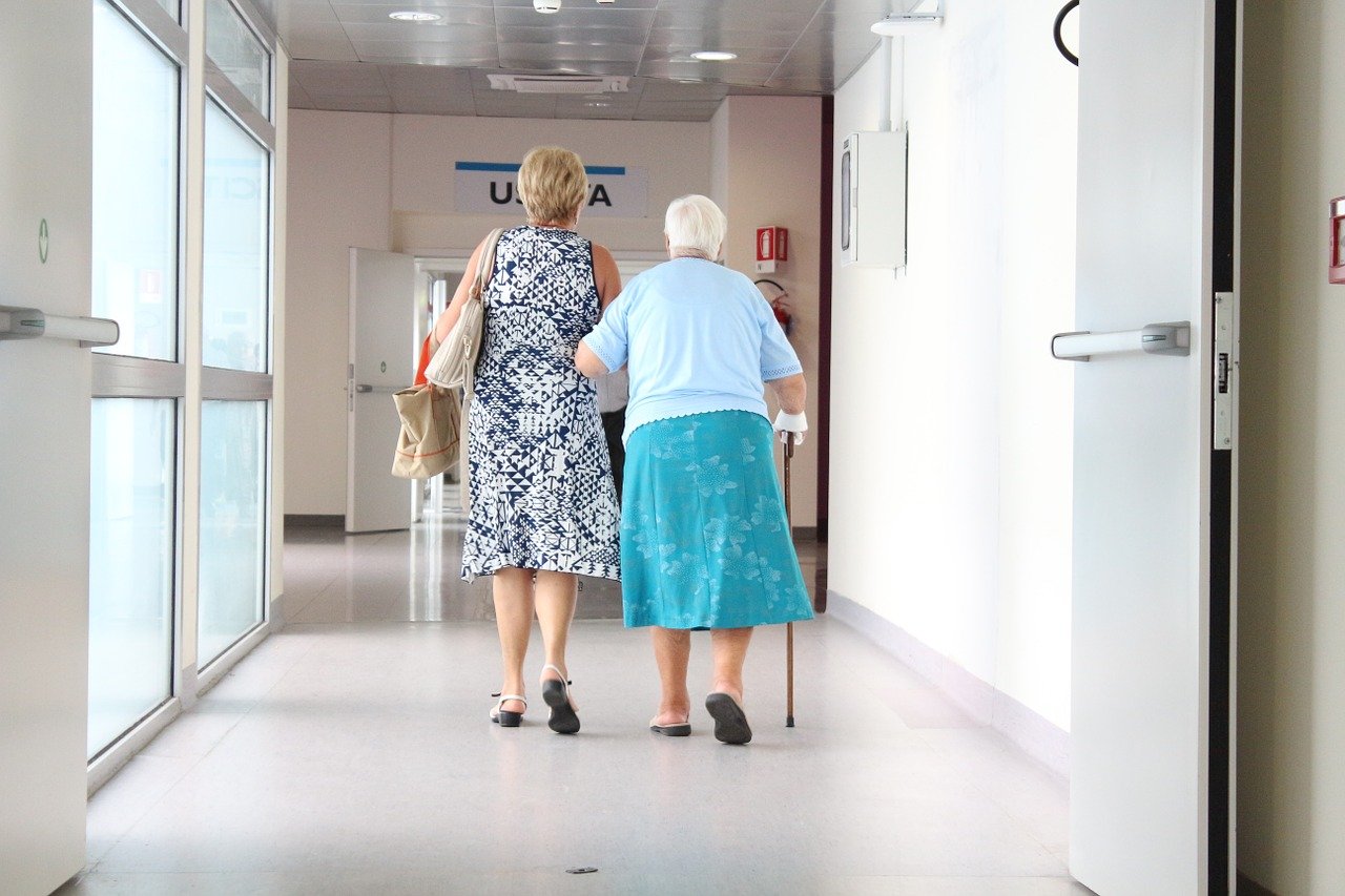 Em um corredor de hospital há duas mulheres de costas. Uma está usando vestido branco com detalhes preto e uma bolsa enquanto acompanha uma idosa vestindo camiseta e saia azul. A idosa está caminhando com uma bengala.