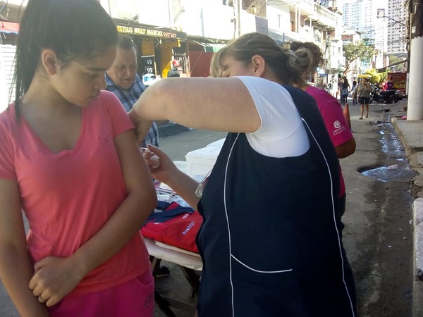 #PraCegoVer: profissional de saúde aplica vacina no braço de uma adolescente, num posto volante montado numa rua de São Paulo.