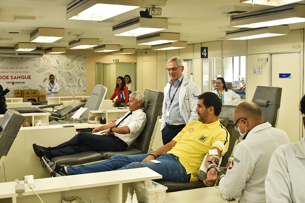 #PraCegoVer: na imagem há dois homens sentados em cadeiras para doação de sangue. No local há pessoas circulando entre os profissionais de saúde e cadeiras de doação de sangue vazias.