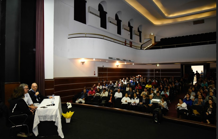 Na imagem feita de cima do palco é possível observar os integrantes da mesa e da plateia cheia durante Encontro de 15 anos de Práticas Integrativas e Complementares em Saúde no Colégio Caetano de Campos