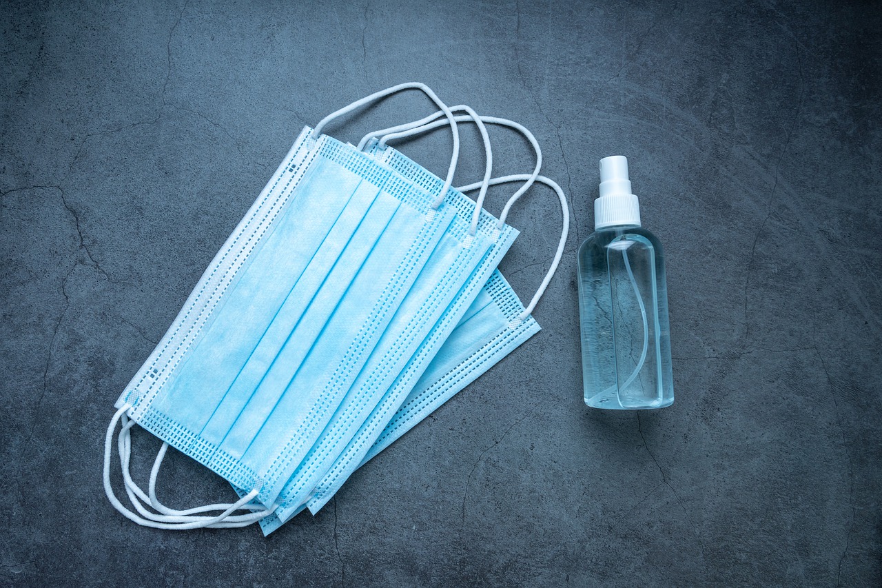 Em um fundo cinza liso, estão quatro máscaras descartáveis nas cores azul e branco, uma em cima da outra. Ao lado delas, está um frasco de álcool em gel transparente, com a tampa de spray branca.