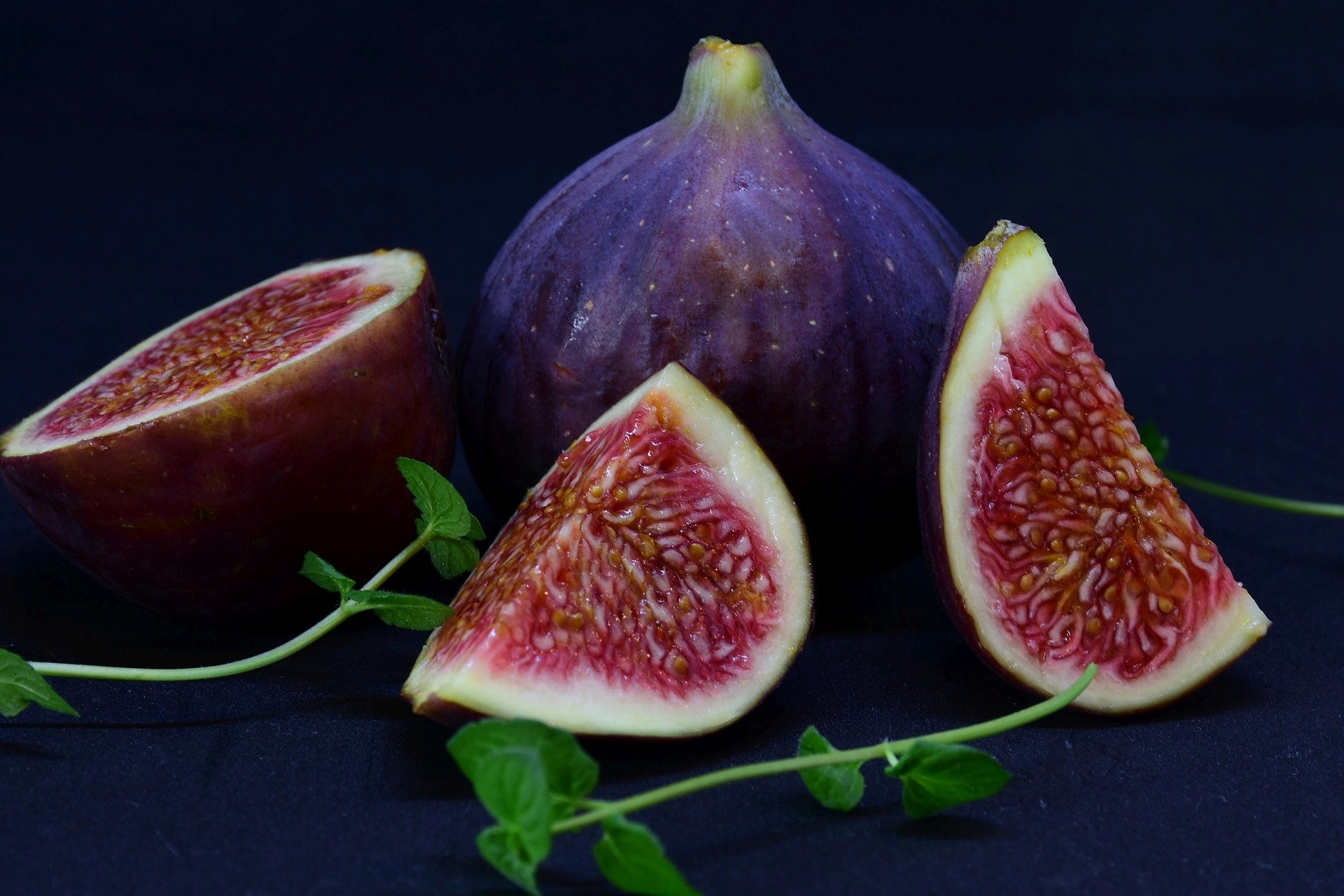 Na imagem há um dois figos, um está inteiro e o outro está cortado em três partes iguais. As frutas estão em cima de uma mesa de pedra preta.