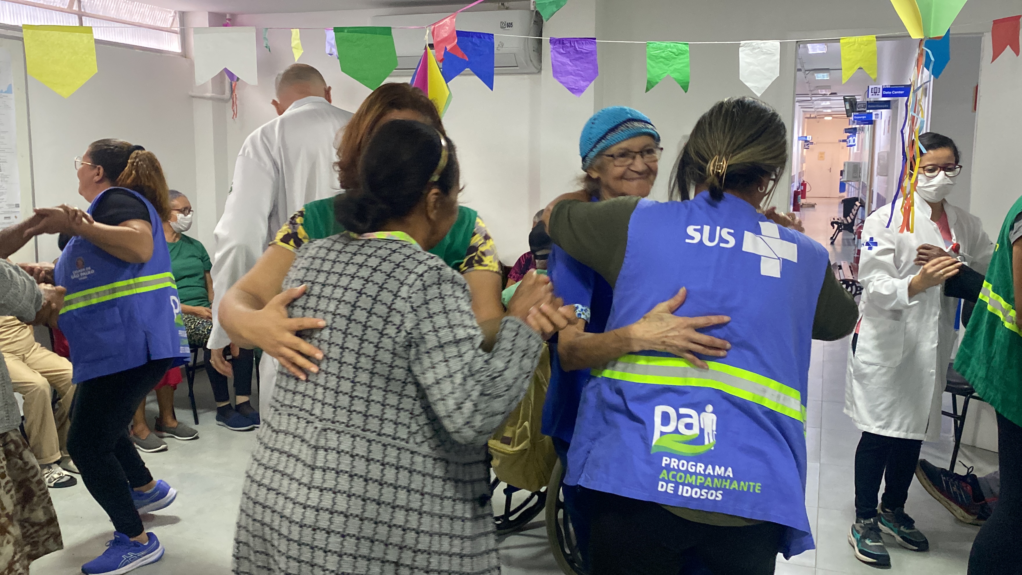 A foto mostra uma sala enfeitada com bandeirinhas de festa junina, onde estão vários idosos, alguns sentados e outros em pé, dançando com profissionais de saúde; duas dessas profissionais usam coletes verdes onde se lê 