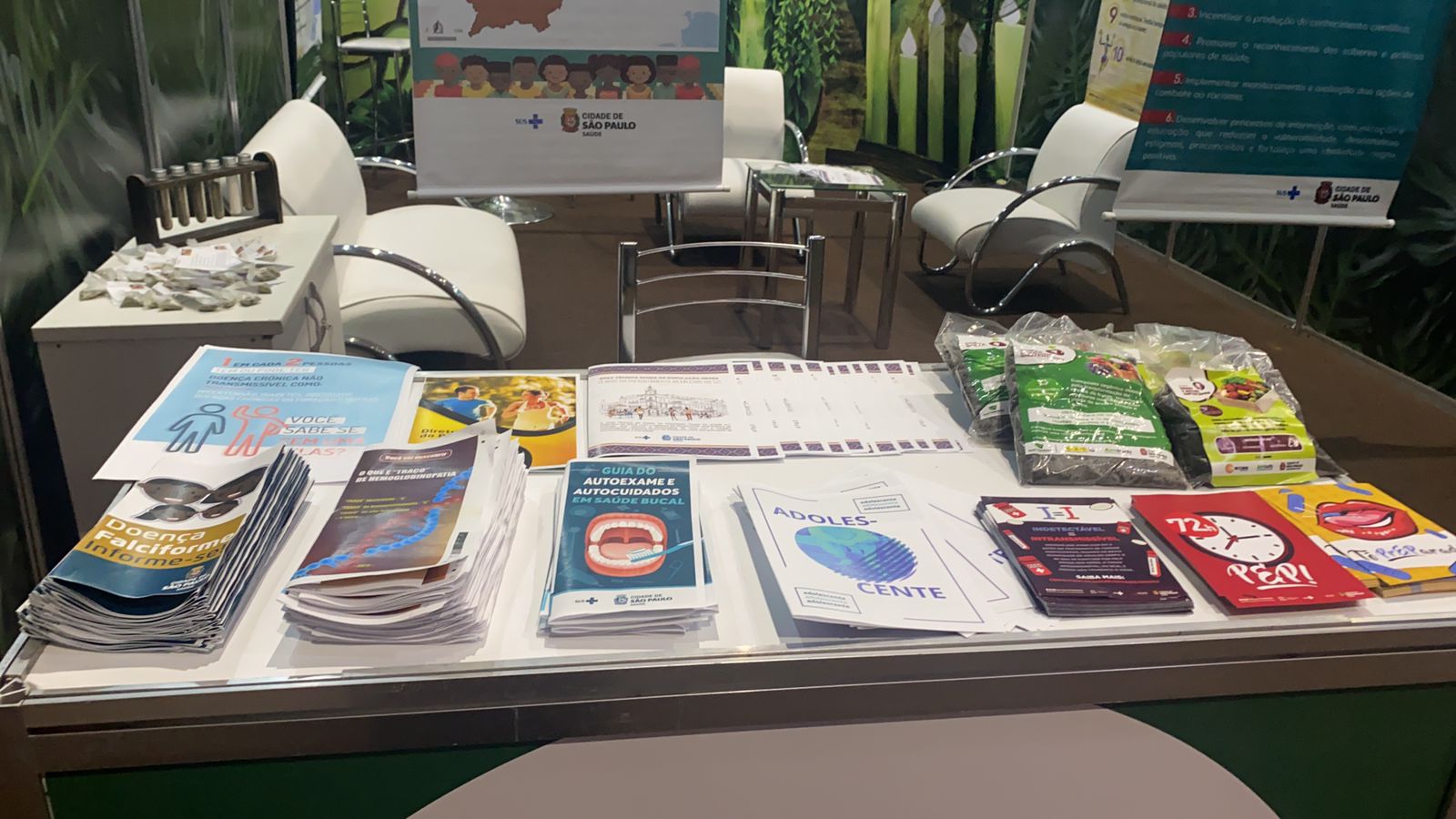 Numa sala com cadeiras com estofados brancos, sobre uma mesa há vários folhetos com temas como: doença falciforme, saúde bucal, adolescente e PEP