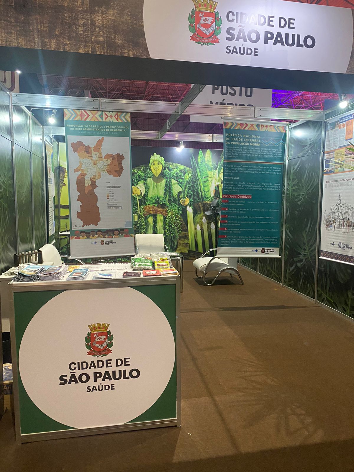 Foto do estande vazio. No primeiro plano, uma mesa com o logotipo da Secretaria Municipal da Saúde e vários folhetos. Ao fundo, três banners com temas diversos.