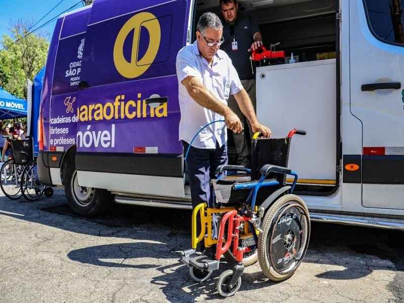 Em frente à uma van que está com a porta aberta, está um homem vestindo camisa branca. Ele está segurando uma cadeira de rodas que está fechada. Na van, azul e branca, está escrito 
