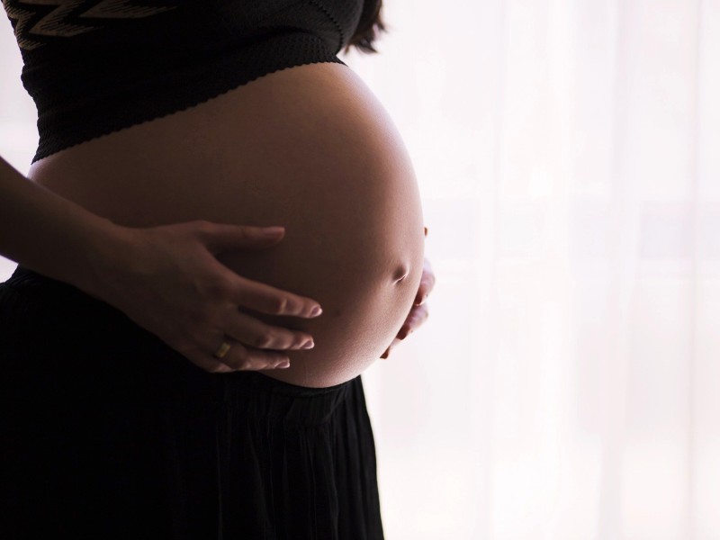 Foto de uma barriga de grávida com fundo branco. A foto foca na barriga de uma mulher. Ela usa saia e blusa curta na cor preta e segura a barriga suavemente com as mãos