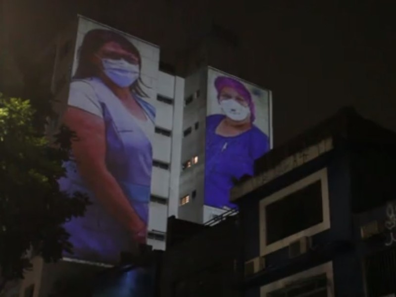 Foto noturna mostra a imagem de duas profissionais de saúde projetadas na lateral de um prédio. Do lado esquerdo há a copa de uma árvore. Também aparecem na foto prédios mais baixos