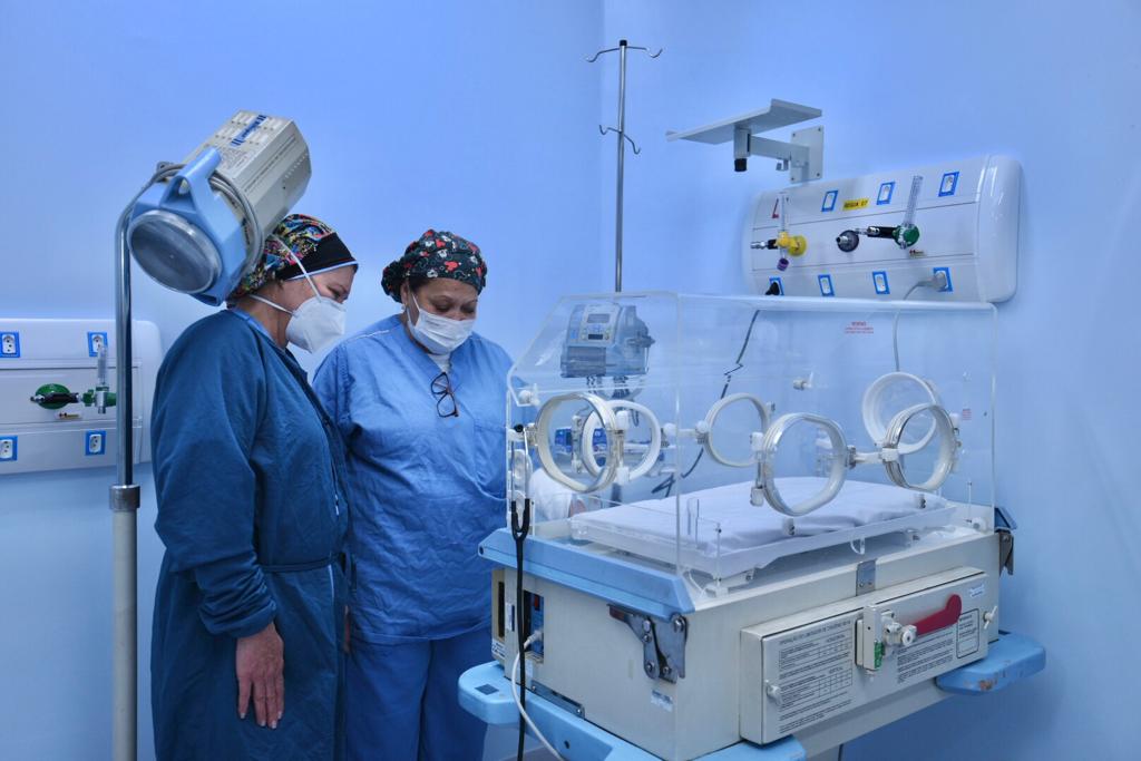 Foto de hospital. Duas profissionais de saúde vestindo aventais azuis, máscaras de proteção brancas e toucas coloridas nos cabelos estão à esquerda da imagem. À direita, está um berço hospitalar vazio. As paredes da sala são azuis.