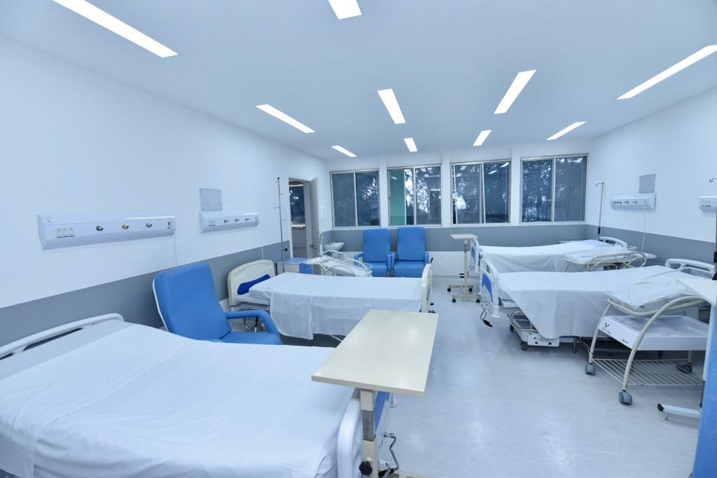 Foto de hospital. As paredes da sala são brancas, com uma faixa cinza do meio ao chão. Ao todo, quatro leitos cobertos com lençóis brancos estão no cômodo. Entre duas camas está uma poltrona azul. Na parede ao fundo da sala, onde estão três janelas de vidro, estão mais duas poltronas azuis.